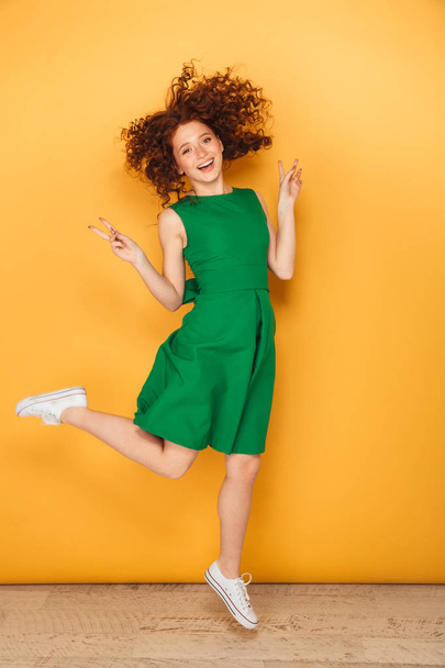 Full length portrait of a cheerful young redhead woman in dress posing while jumping isolated over yellow background - 写真・画像