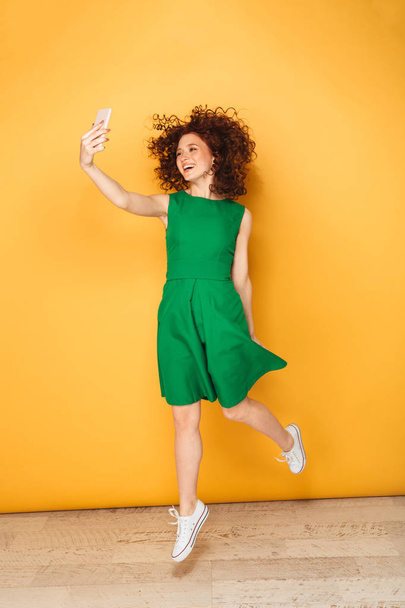 Full length portrait of a cheerful redhead woman in dress taking a selfie isolated over yellow background - Φωτογραφία, εικόνα