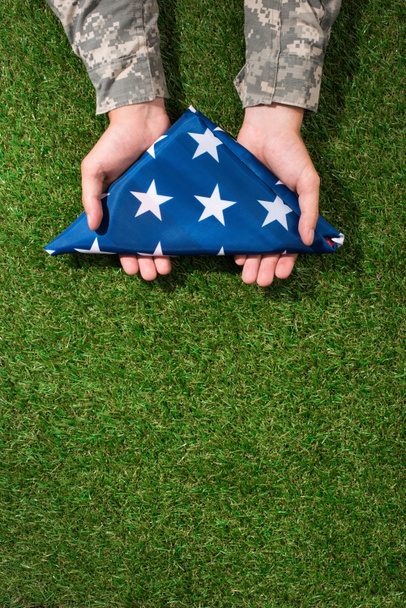 częściowy widok żołnierz w mundurze wojskowym gospodarstwa złożone flaga w ręce na zielonej trawie, 4 lipca wakacje koncepcja - Zdjęcie, obraz