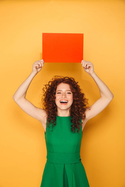 Portrait of a joyful redhead woman in dress holding empty copy space isolated over yellow background - Foto, Bild