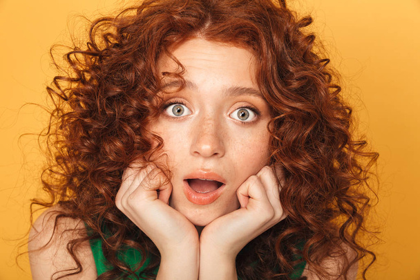 Close up portrait of a surprised curly redhead woman looking at camera isolated over yellow background - 写真・画像