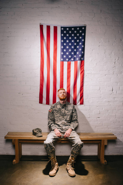 red hair american soldier sitting on wooden bench with flag on white brick wall behind, americas independence day concept - Photo, Image