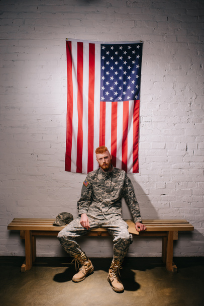 soldado en uniforme militar con carta sentado en banco de madera con bandera americana en la pared de ladrillo blanco detrás, 4 julio concepto de vacaciones
 - Foto, Imagen