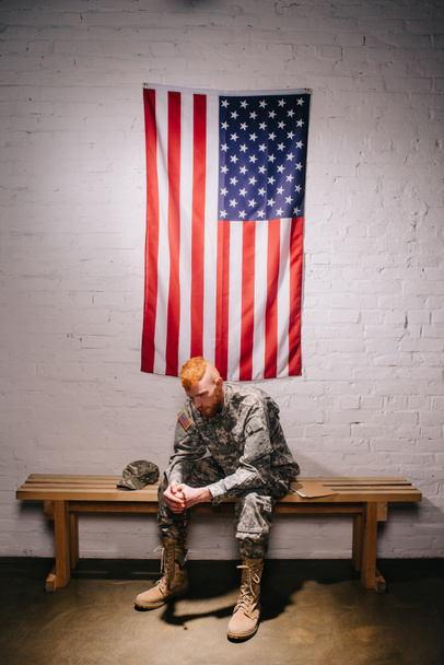 soldado em uniforme militar sentado em banco de madeira com bandeira americana na parede de tijolo branco atrás, conceito de feriado de 4 de julho
 - Foto, Imagem