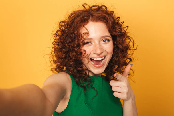 Close up portrait of a cheerful redhead woman in dress taking a selfie and pointing finger isolated over yellow background - Foto, imagen