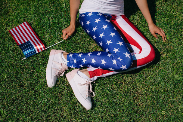 vista parcial de la mujer con asta de la bandera en leggins con patrón de bandera americana descansando en el césped verde, concepto de vacaciones del día de independencia de América
 - Foto, Imagen