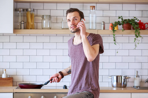 Photo of brunet man with frying pan in his hands talking on phone in kitchen - Photo, Image