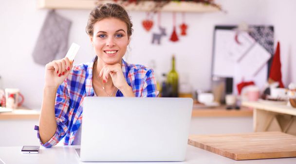 Mujer sonriente compras en línea utilizando la computadora y la tarjeta de crédito en la cocina
 - Foto, imagen