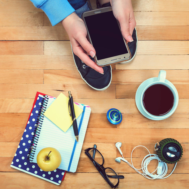 Female hand with a mobile phone, notebooks, pen, glasses, music speakers, headphones, cup of tea, apple on a wooden background - Photo, Image