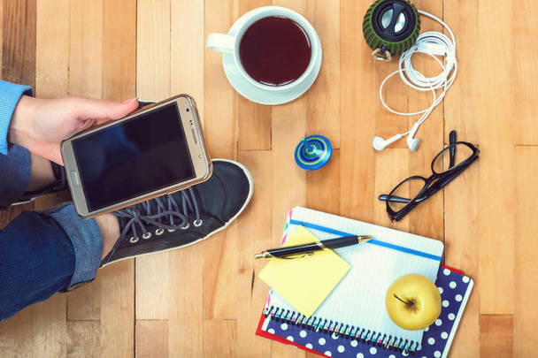 Ženská ruka s mobilní telefon, poznámkové bloky, pero, brýle, hudební reproduktory, sluchátka, čaj, jablko na dřevěné pozadí - Fotografie, Obrázek