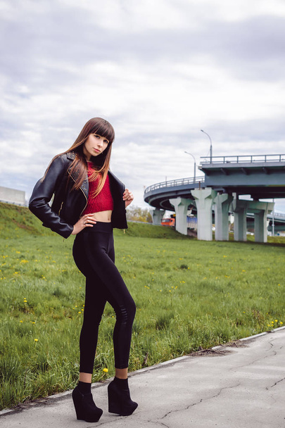 young beautiful stylish woman in fashionable black clothes, a burgundy cardigan and a dark leather jacket posing outdoors near the main road junction - Фото, зображення