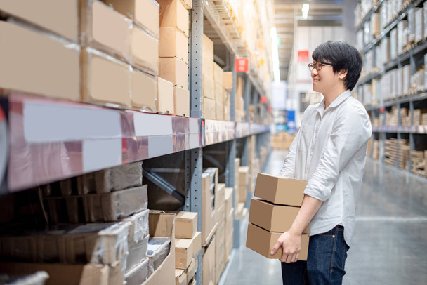 Young Asian man carrying cardboard boxes near product shelf in warehouse, shopping warehousing or working pick and packing concepts - Photo, Image