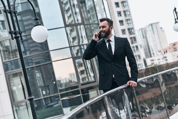 handsome businessman in suit talking on phone while standing outdoors - Foto, Bild