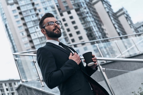 thoughtful businessman holding disposable cup with coffee and looking away while standing outdoors - Foto, afbeelding