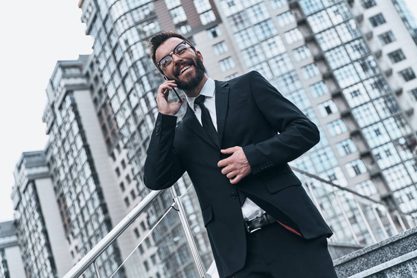 smiling businessman in suit talking on mobile at buildings  - Фото, изображение
