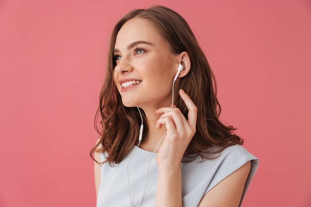 Image of cheerful young woman standing isolated over pink background listening music with earphones. Looking aside. - Fotoğraf, Görsel