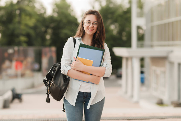 Portrait of a student girl with glasses. She stands in the background of the park and holds notebooks and folders in her hands. The girl is preparing to enter the university - Foto, Imagem