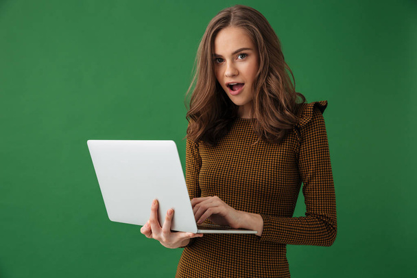 Image of young emotional woman standing isolated over green background holding laptop computer looking camera. - Fotó, kép