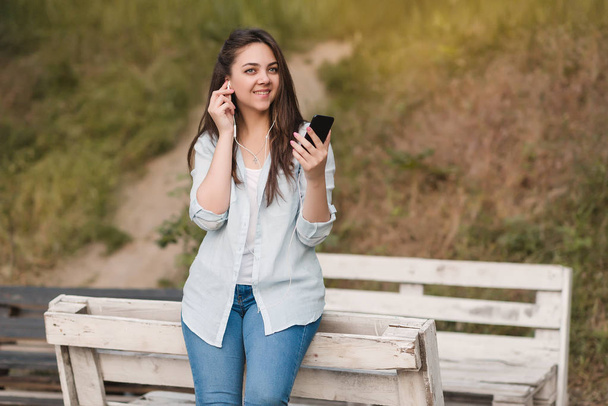 Een meisje zit op een houten bankje in het park en luistert met vrolijke muziek op koptelefoon - Foto, afbeelding
