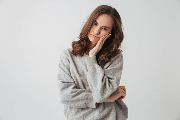 Upset brunette woman in sweater touching her cheek while looking at the camera over grey background - Foto, Imagem