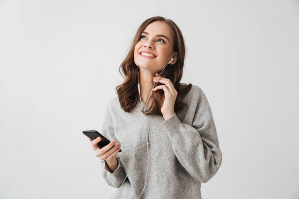 Pensive smiling brunette woman in sweater holding smartphone and looking up over grey background - Foto, Imagen