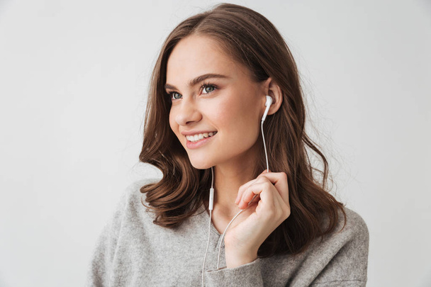 Smiling brunette woman in sweater listening music by earphones while looking away over grey background - Photo, image