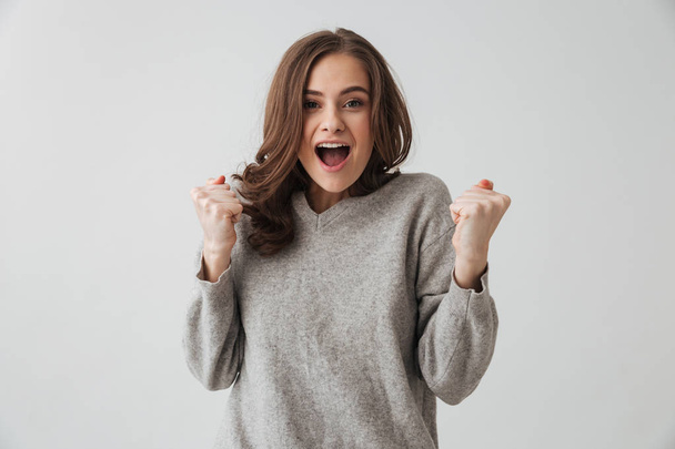 Surprised happy brunette woman in sweater rejoices and looking at the camera over grey background - Valokuva, kuva