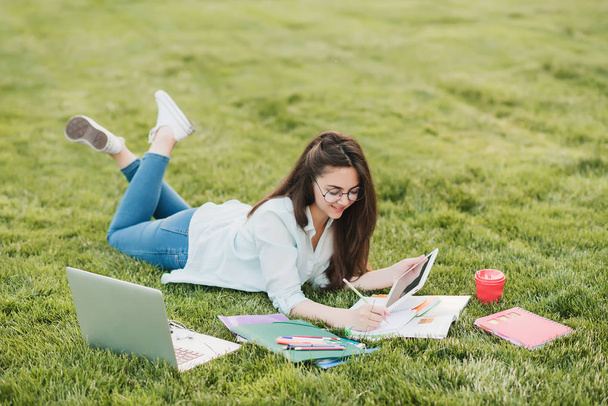 A young student lies on a green lawn in front of the university and is preparing for the final exams. She scans the scientific articles in the Internet and makes useful notes in the notebook - Fotó, kép