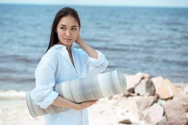 attractive asian woman holding yoga mat and looking away by sea  - Photo, Image
