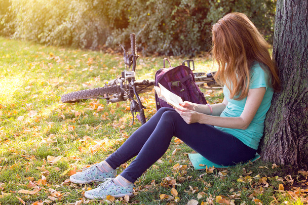 公園で少女は、ツリーの下に座っているし、本を読みます。その隣には自転車と、backpac です。 - 写真・画像