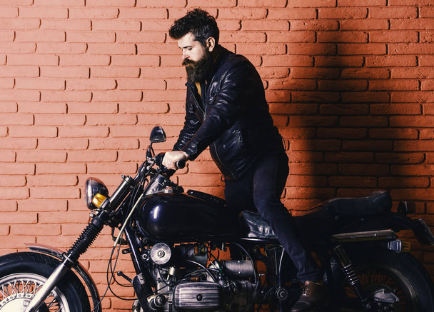brutal biker on serious face in leather jacket gets on motorcycle. Man with beard, biker in leather jacket near motor bike in garage, brick wall background. Start of journey concept - Foto, afbeelding