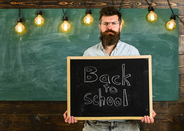 Teacher in eyeglasses holds blackboard with inscription back to school. Man with beard and mustache on strict face welcomes students, chalkboard on background. Back to school concept - Foto, imagen