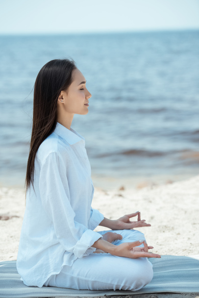 vista lateral da jovem mulher asiática em ardha padmasana (meia postura de lótus) no tapete de ioga por mar
  - Foto, Imagem