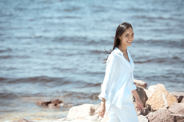 happy young asian woman looking at camera while standing on beach by sea  - Фото, зображення
