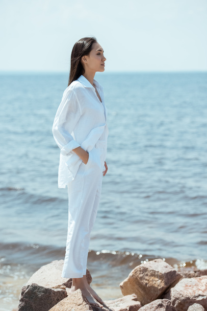 side view of attractive asian woman with hand in pocket standing on beach by sea  - Foto, Imagem