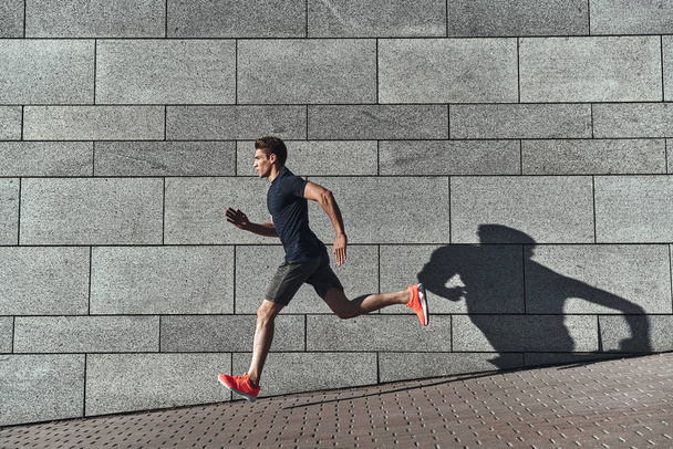 handsome young man in sports clothing running in city street at stone wall - Fotoğraf, Görsel