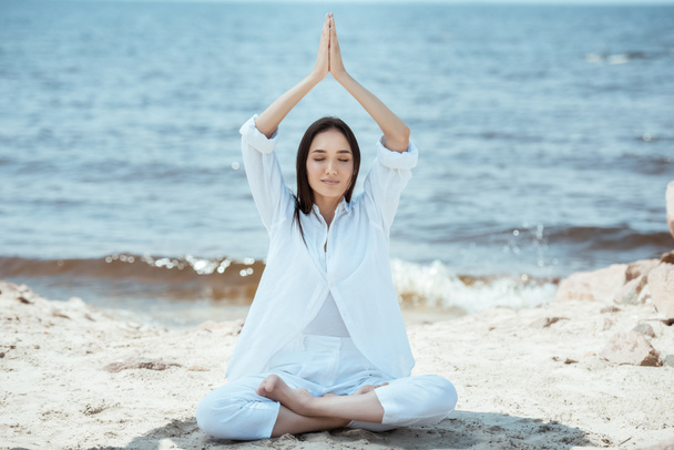 fokussierte asiatische junge Frau meditiert in Lotusposition und macht namaste mudra Geste über Kopf am Meer  - Foto, Bild