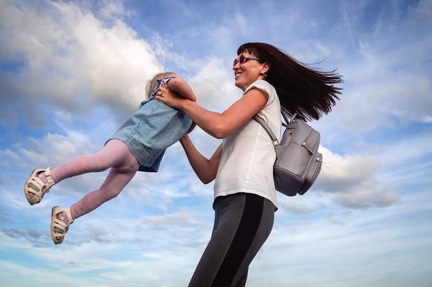 Family values: Mom with sunglasses whirl with a little daughter in a blue dress against the sky and clouds. Authentic family. - 写真・画像