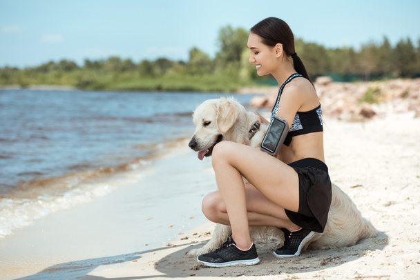 happy asian sportswoman with smartphone in running armband case embracing golden retriever on beach  - Fotografie, Obrázek