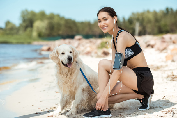 smiling asian sportswoman in earphones with smartphone in running armband case tying shoelaces near golden retriever on beach  - Photo, Image