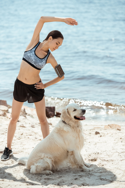 asian sportswoman with smartphone in running armband case doing exercise while her dog sitting near on beach  - Photo, Image