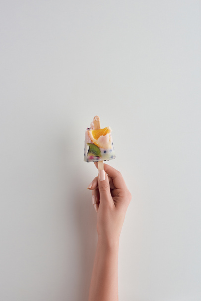 περικοπεί shot του προσώπου που κρατάει φρέσκο κρύο φρουτώδες popsicle στο stick σε γκρι  - Φωτογραφία, εικόνα