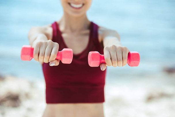 cropped image of smiling sportswoman exercising with dumbbells in front of sea - Фото, изображение