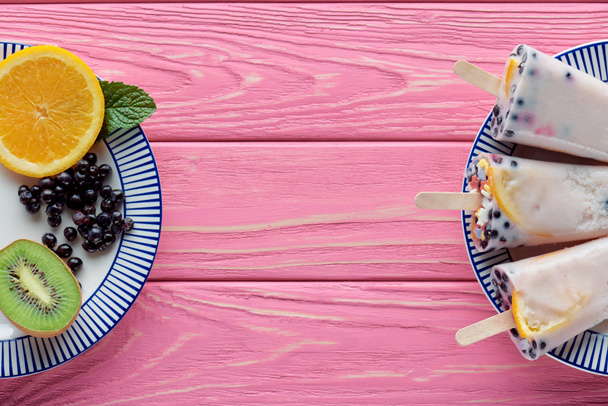 top view of homemade popsicles and fresh fruits on plates on pink wooden table - Foto, afbeelding