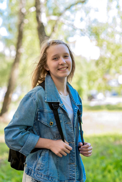 Happy teen girl smiling. Summer in nature. A schoolgirl on vacation. In denim jacket behind a backpack. Emotions of love and happiness. - Foto, imagen