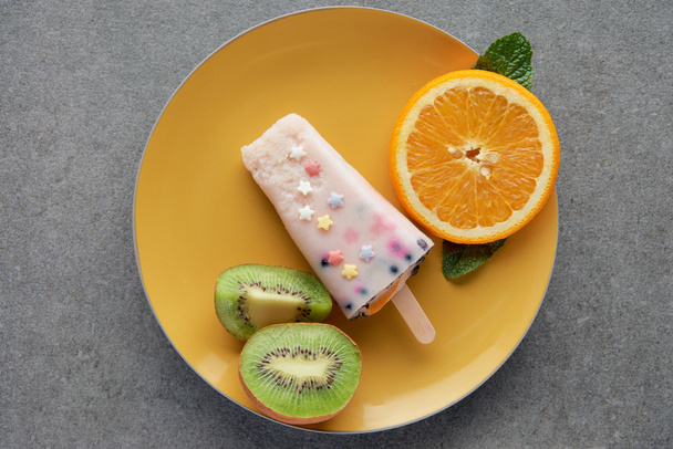 top view of delicious popsicle with slices of orange and kiwi on yellow plate on grey - 写真・画像