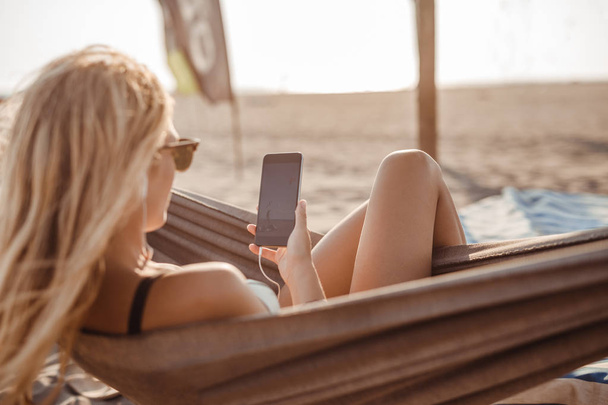 Pretty Caucasian woman lying on hammock at beach and typing on her cell phone. - Foto, Imagem
