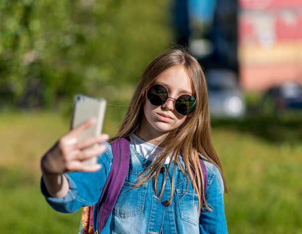 Little girl schoolgirl. Summer in nature. In his hands holds a smartphone. In sunglasses and a denim jacket. He takes pictures of himself on a smartphone. Make a video call over the Internet. - Photo, image