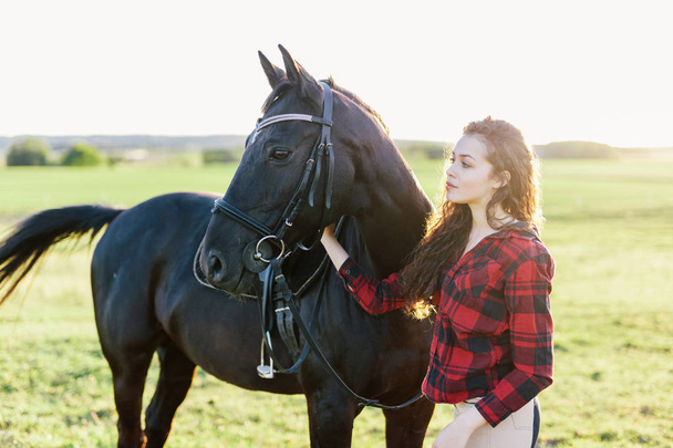 Musta hevonen ja viehättävä nuori nainen seisoo kentällä
. - Valokuva, kuva
