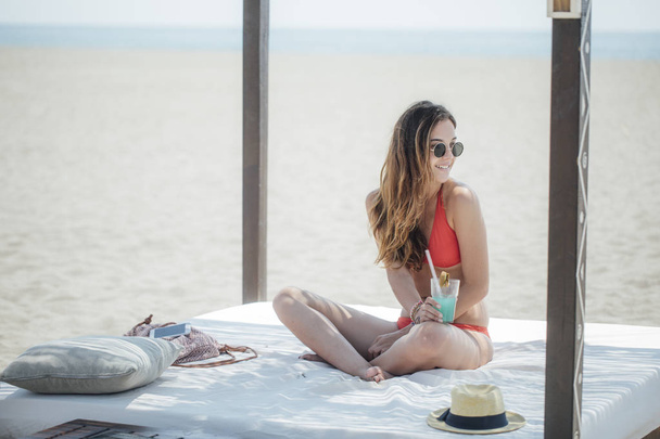 Young Caucasian smiling girl sitting on baldachin on sandy beach and drinking cocktail. - Foto, Imagen